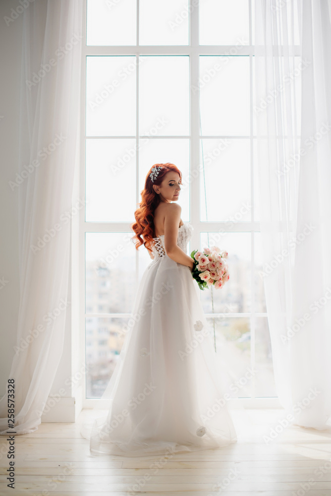 Red-haired bride in white wedding dress stands near large window Red-haired bride in white wedding dress stands near large window