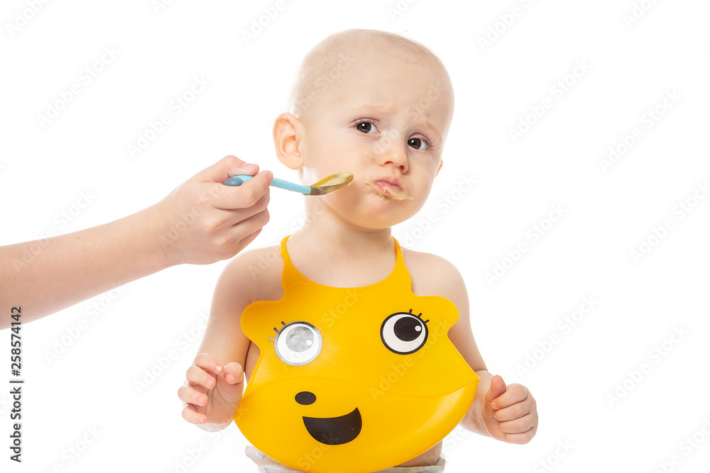 Baby boy does not want to eat porridge spoon mashed. Infant baby posing in yellow bib Isolated on white background. family, food, eating and people concept Baby boy does not want to eat porridge spoon mashed. Infant baby posing in yellow bib Isolated on white background. family, food, eating and people concept