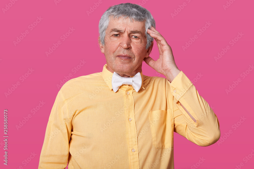 Φωτογραφία Studio shot of handsome old man keeps hand on temple, has headache, wears yellow shirt and white bow tie, has embrassed facial expression, isolated over pink studio background