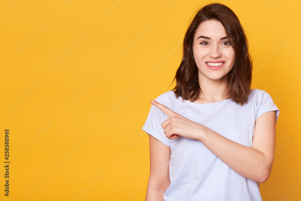 Portrait of cute charming cheerful girl pointing aside with index finger, wears white casual t shirt, has dark hair, isolated over yellow background. Copy space for your promotion text or advertisment Portrait of cute charming cheerful girl pointing aside with index finger, wears white casual t shirt, has dark hair, isolated over yellow background. Copy space for your promotion text or advertisment