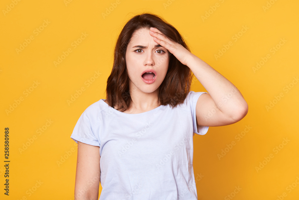 Portrait of forgetful female model remembers something, keeps hand on forehead, opens mouth with astonishment, remembers to do importent task, wears white casual t shirt. Bad memory concept. Portrait of forgetful female model remembers something, keeps hand on forehead, opens mouth with astonishment, remembers to do importent task, wears white casual t shirt. Bad memory concept.