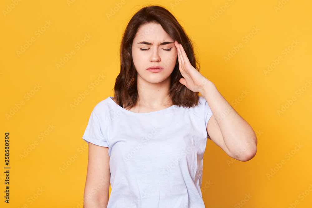 Close up portrasit of young brunette woman wearing white casual t shirt isolated over yellow background with hand on temple, has pain in head because stress, stands with closed eyes. Copy space.