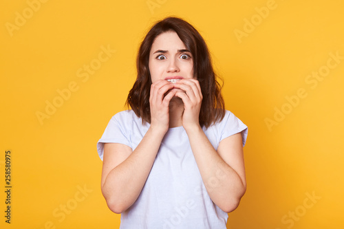 Portrait of frightened brunette girl bites fingers nails, has frightneed facial expression, afraids of something, wears in casual white t shirt, models on yellow background. Negative feelings concept
