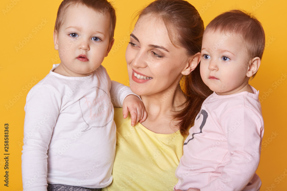 Portrait of young mother holding her little twins, cute active infants being interested to be photografed, look asides, mother looks with love on her adorable daughters. happy family concept. Portrait of young mother holding her little twins, cute active infants being interested to be photografed, look asides, mother looks with love on her adorable daughters. happy family concept.