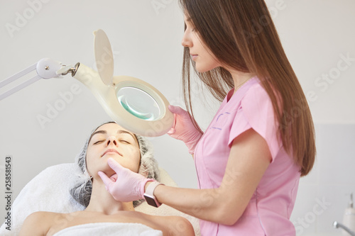 Cosmetologist dresses pink madical gown, latex gloves examines patient face, finds defects under lamp, prepering woman for ejuvenation and hydration procedures. Cosmetology, aethetic medicine concept.