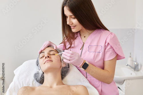 Cosmetologist wears pink medical gown makes beauty tretment for female patient in beauty salon, applys hyaluronic acid. Facial Treatment, essence skin care, cosmetic, face rejuvenation concept. photo