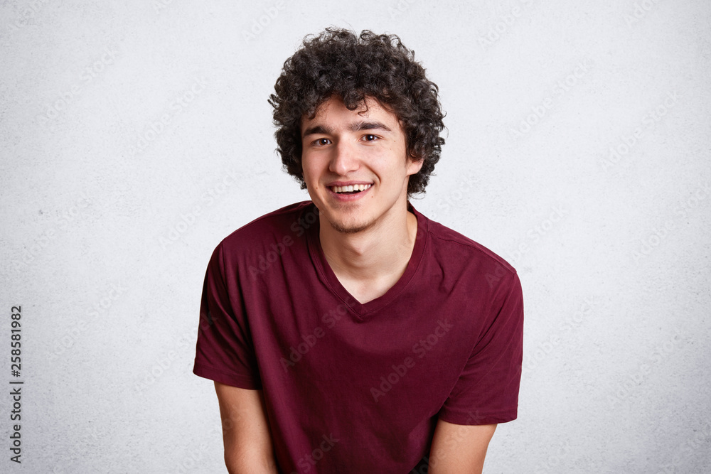 Horizontal shot of happy handsome male with shining smile, feels glad, has specific appearance, dressed in fashionable maroon t shirt, poses isolated over white background. Copy space for promotion. Horizontal shot of happy handsome male with shining smile, feels glad, has specific appearance, dressed in fashionable maroon t shirt, poses isolated over white background. Copy space for promotion.