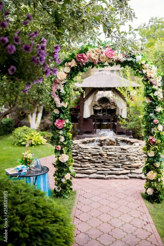 Fototapeta Naklejka Na Ścianę i Meble -  Romantic place with floral arch and fountain made in stones in the garden. Arch for the wedding ceremony, decorated with flowers over the natural background.