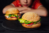 Two delicious burgers with tomato, beef, green cucumber and lettuce on the background of a boy. Sad boy sitting at the table and looking at tasty hamburgers.