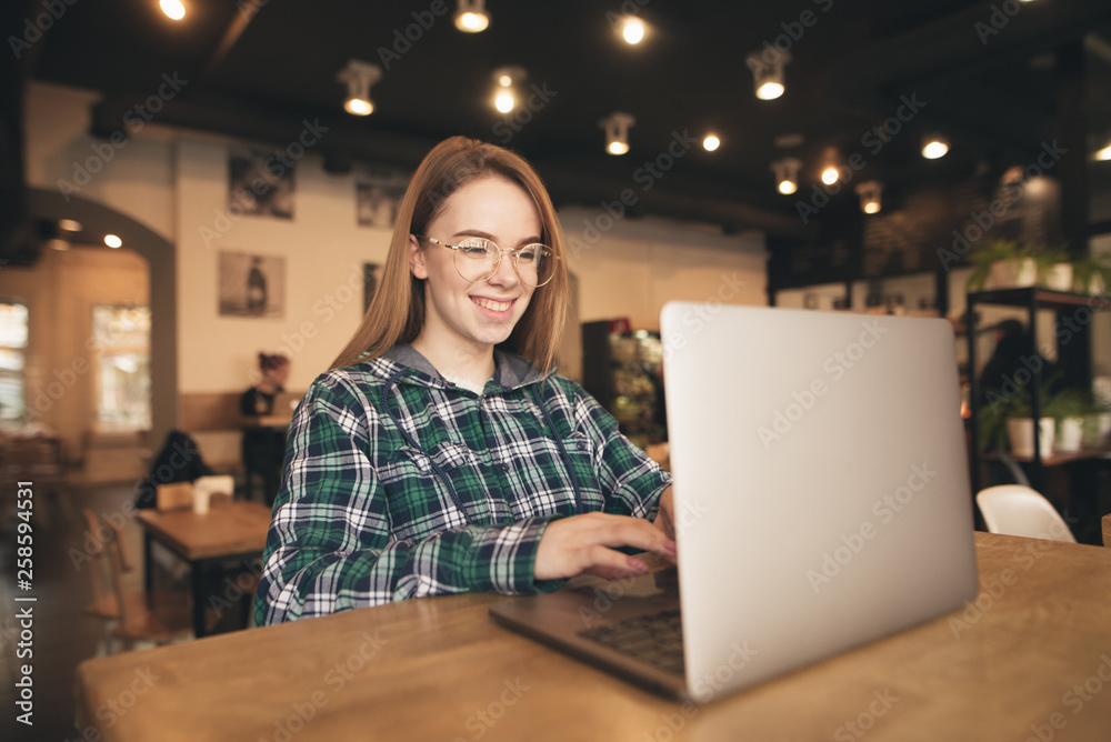 Portrait of a happy freelancer sits in a cozy cafe with a laptop, dials text on the keyboard and smiles. Positive blogger girl works in a cafe with a laptop. Student studying in a cafe. Portrait of a happy freelancer sits in a cozy cafe with a laptop, dials text on the keyboard and smiles. Positive blogger girl works in a cafe with a laptop. Student studying in a cafe.