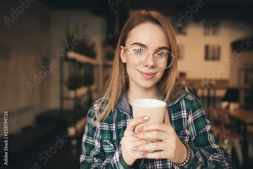 Cute girl with a cup of coffee in her hands in a cozy coffee shop, wearing a shirt and casual clothing, looks at the camera and smiles. Close up portrait