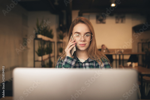 Portrait of a beautiful girl in casual clothes and glasses talking on the phone and using a laptop in a cozy cafe. Student girl works in a cafe.