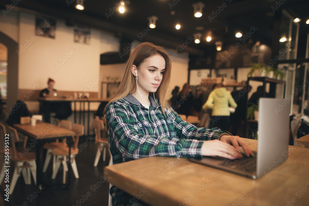 Portrait of a stylish girl wearing a shirt, using a laptop in the coffee shop, looking at the screen and listening to the sound in the headphones. Beautiful girl works on a laptop in a cafe.