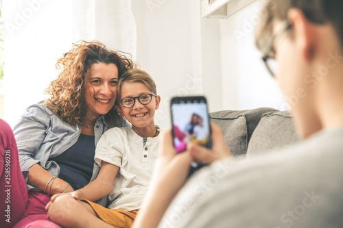 Young boy takes picture of his mother and son sitting on a sofa at home. Smiling family play with smartphone Mum and son enjoy a day off with older brother Familiar scene of happy people taking selfie