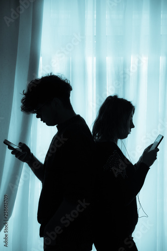 Silhouette of two teenagers with cell phones standing back to back, cold effect