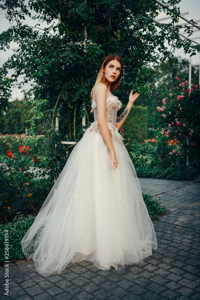 Young beautiful model is posing in a long ivory dress in the garden with a crown on her head Young beautiful model is posing in a long ivory dress in the garden with a crown on her head