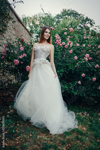 Young beautiful model is posing in a long ivory dress in the garden with a crown on her head 