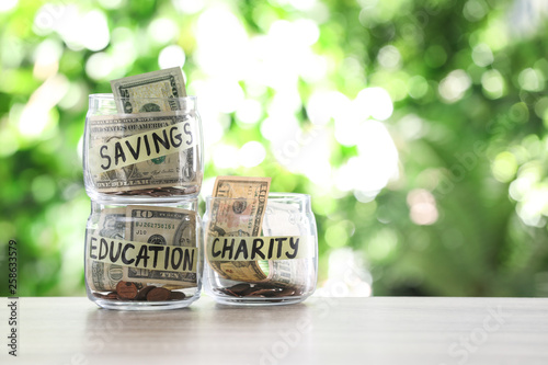 Glass jars with money for different needs on table against blurred background