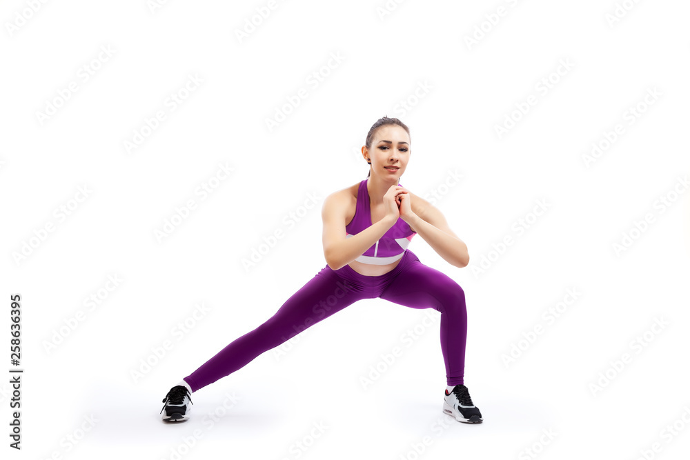 A young woman coach in a sporty pink   short top and gym leggings makes lunges  by the feet side, hands are held out to the side   on a  white isolated background in studio