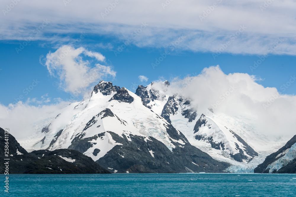 Snow and glacier ice covered rocky mountain peaks, with low white clouds, glacier melt blue water, and blue sky and white clouds above, Drygalski Fjord, South Georgia