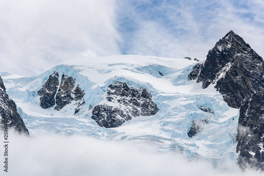 Snow and glacier ice covered rocky mountain peak, with fog below and light white clouds in a blue sky above, Drygalski Fjord, South Georgia