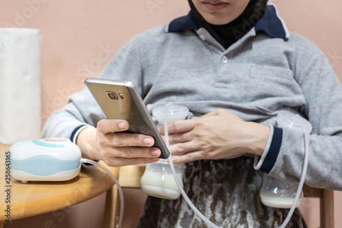 Busy mother pumping breastmilk with automatic breast pump machine while using mobile phone.