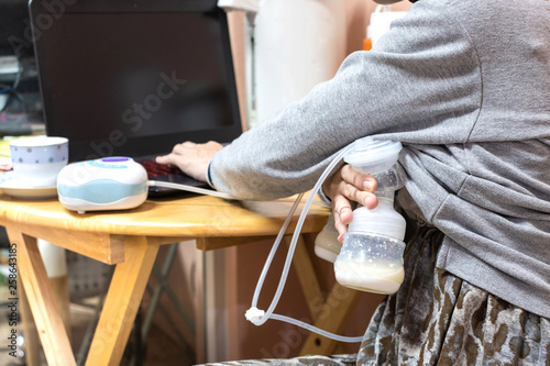 Busy mother pumping breastmilk with automatic breast pump machine while using laptop.