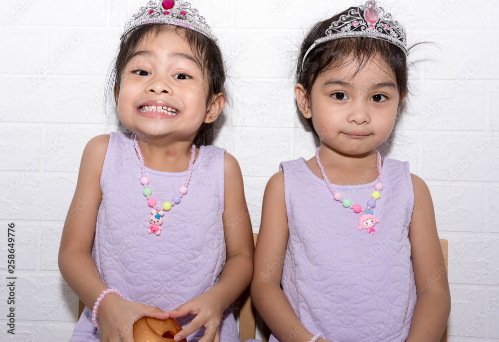Female asian identical twins sitting on chair with white background. Wearing purple dress and accessories like necklace, crown Female asian identical twins sitting on chair with white background. Wearing purple dress and accessories like necklace, crown