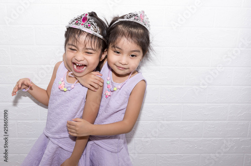Female asian identical twins sitting on chair with white background. Wearing purple dress and accessories. Standing hugging each other