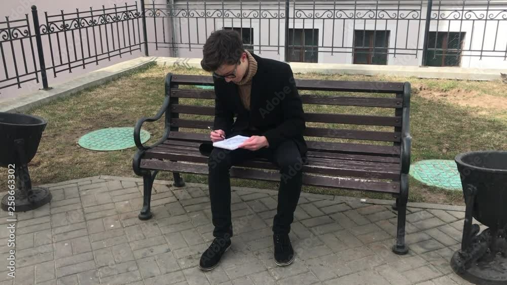 A young man sits on a bench and makes sketches in a notebook. Spring, cool weather, the guy is wearing a black coat. Cozy courtyard in retro style.. A young man sits on a bench and makes sketches in a notebook. Spring, cool weather, the guy is wearing a black coat. Cozy courtyard in retro style..