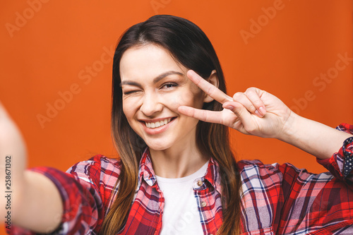 Portrait of pretty young happy woman making selfie on smartphone, isolated against orange background.