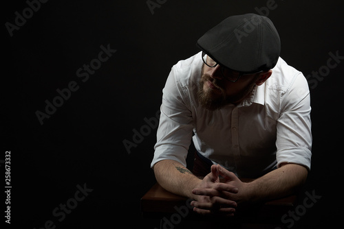young handsome man in the glasses of struggle and mustache in a black cap and a white shirt conceived bent on his hands, a dark studio background