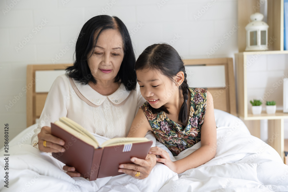 grandmother and granddaughter sitting and read book on the bedroom and white bed
