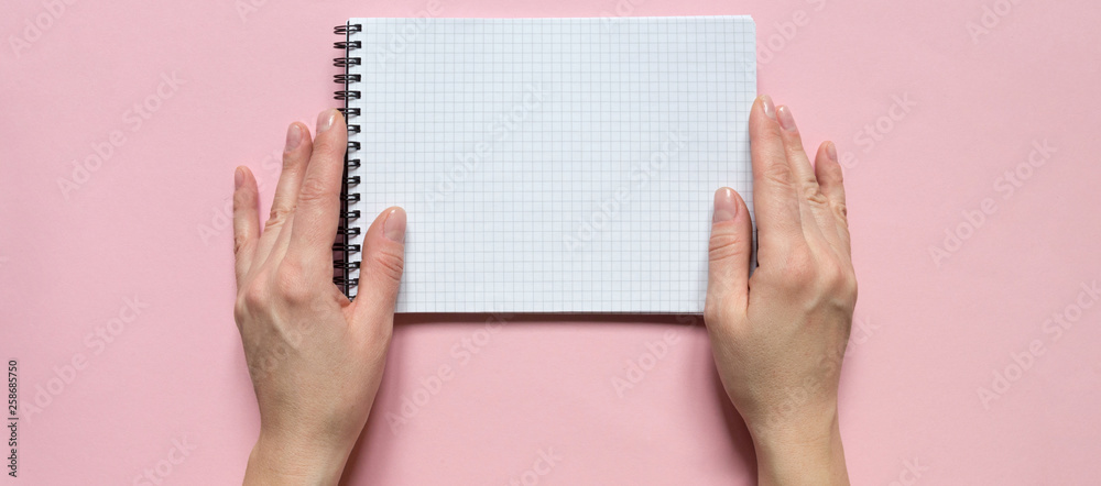 Female hands holding a notebook with blank sheets for writing on a pink background. Flat lay top view. Female hands holding a notebook with blank sheets for writing on a pink background. Flat lay top view.