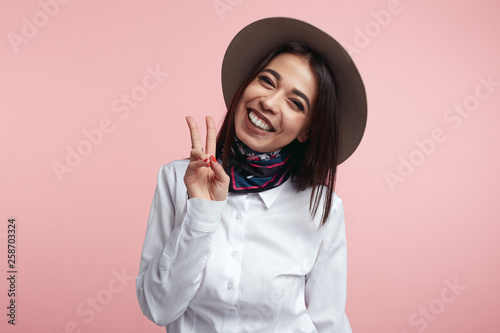 Pleased attractive woman shows v sign or peace gesture, wears white shirt and hat, isolated over rosy studio background. People, fashion, body language.