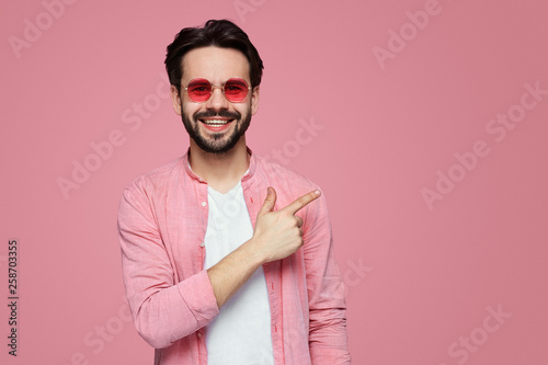 Stylish unshaven man in stylish shirt at copy space on pink wall points aside with cheerful expression, shows something amazing at blank space, isolated over bright pink backgroud © Davidovici