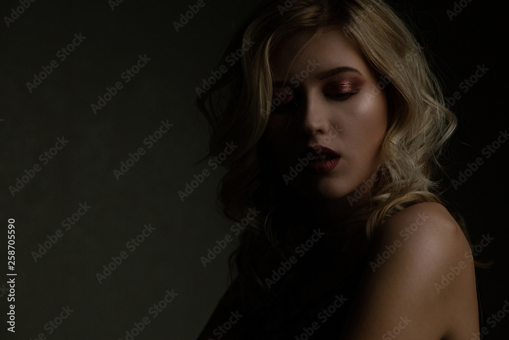 Tender blonde girl with perfect makeup and naked shoulders posing with contrast light. Copy space