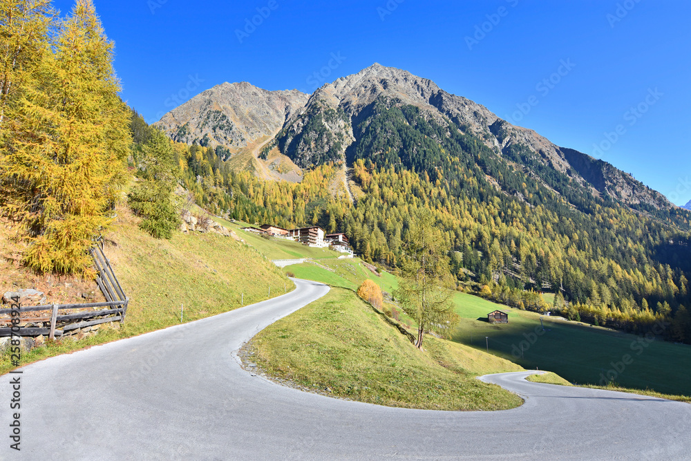 Winding road towards the small hamlet Winnebach near the Oetztal valley (Stubai Alps, Tyrol, Austria). Houses are surrounded by colorful autumn forest and rocky mountains under blue sky.
