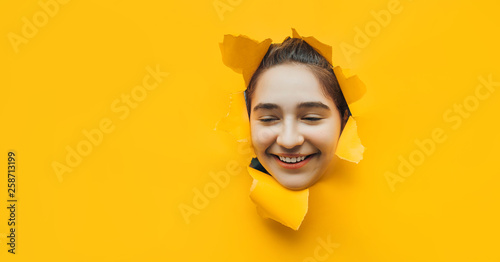 Laughing funny teenage caucasian girl peeping through hole on yellow paper. The concept of surprise, joyful mood from what he saw. Discounts, sales, surprise, smile. Copy space.