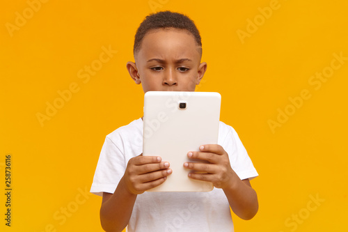 Isolated image of serious concentrated African little boy holding generic touchpad tablet using wireless high speed internet connection, playing video games online, having focused facial expression
