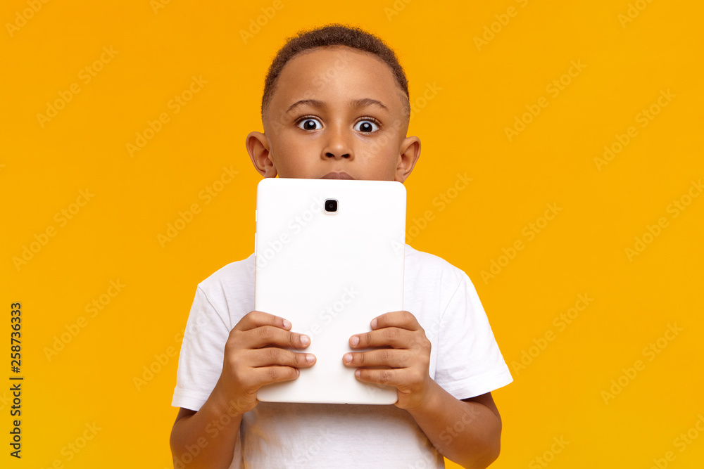 People, children, modern lifestyle, technology and communication concept. Emotional bug eyed dark skinned schoolboy holding digital tablet at his face, staring at camera with astonished look People, children, modern lifestyle, technology and communication concept. Emotional bug eyed dark skinned schoolboy holding digital tablet at his face, staring at camera with astonished look