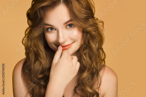 Portrait of nice young woman with long red wavy silky hair, natural make up smiling on camera with hand near lips. Isolated on yellow background. People emotions and feelings. Beauty concept