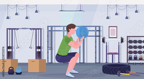 sporty man doing squats exercises with medicine leather ball guy training cardio workout concept modern gym health studio club interior horizontal full length