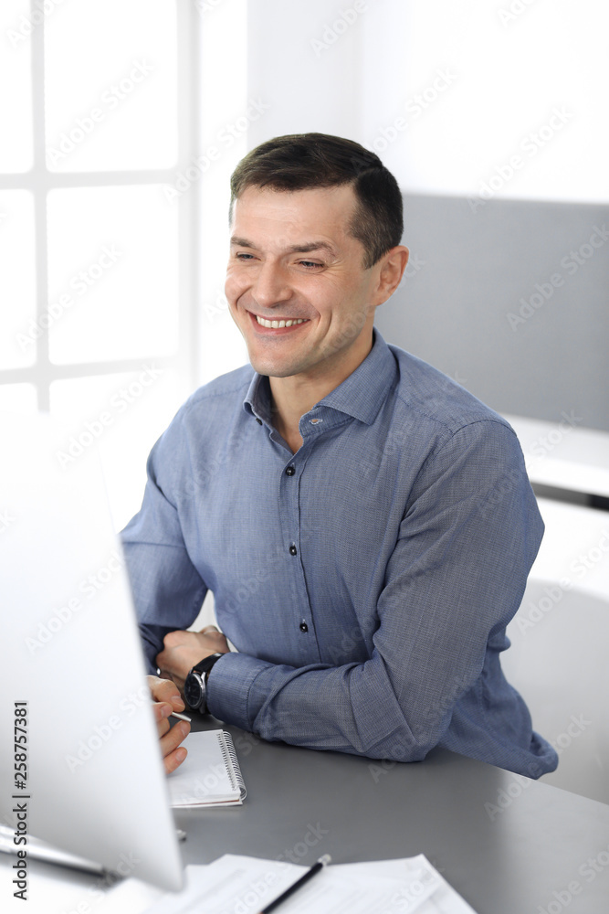 Naklejka premium Cheerful smiling businessman working with computer in modern office. Headshot of male entrepreneur or director of a company at the workplace. Business concept 