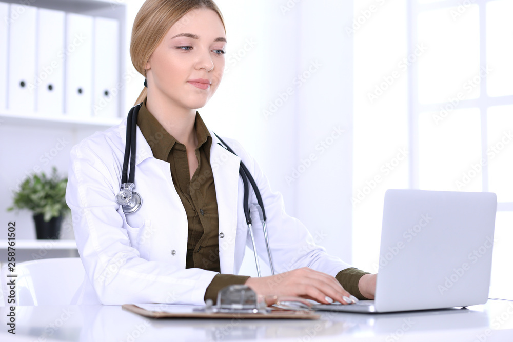 Young woman doctor at work while typing on laptop computer in hospital office. Physician controls medication history records and exam results. Medicine and healthcare concept Young woman doctor at work while typing on laptop computer in hospital office. Physician controls medication history records and exam results. Medicine and healthcare concept