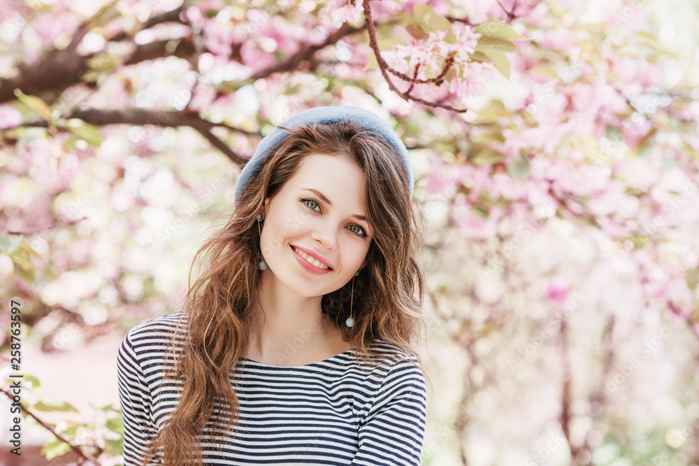 Outdoor close up spring portrait of young beautiful fashionable happy smiling lady with long hair posing in street with blooming pink sakura trees on background. Copy, empty space for text Outdoor close up spring portrait of young beautiful fashionable happy smiling lady with long hair posing in street with blooming pink sakura trees on background. Copy, empty space for text
