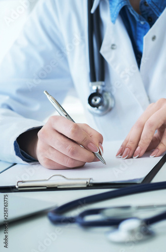 Female doctor filling up prescription form or patient history list at clipboard pad during physical exam or disease prevention while sitting at the desk in hospital closeup.