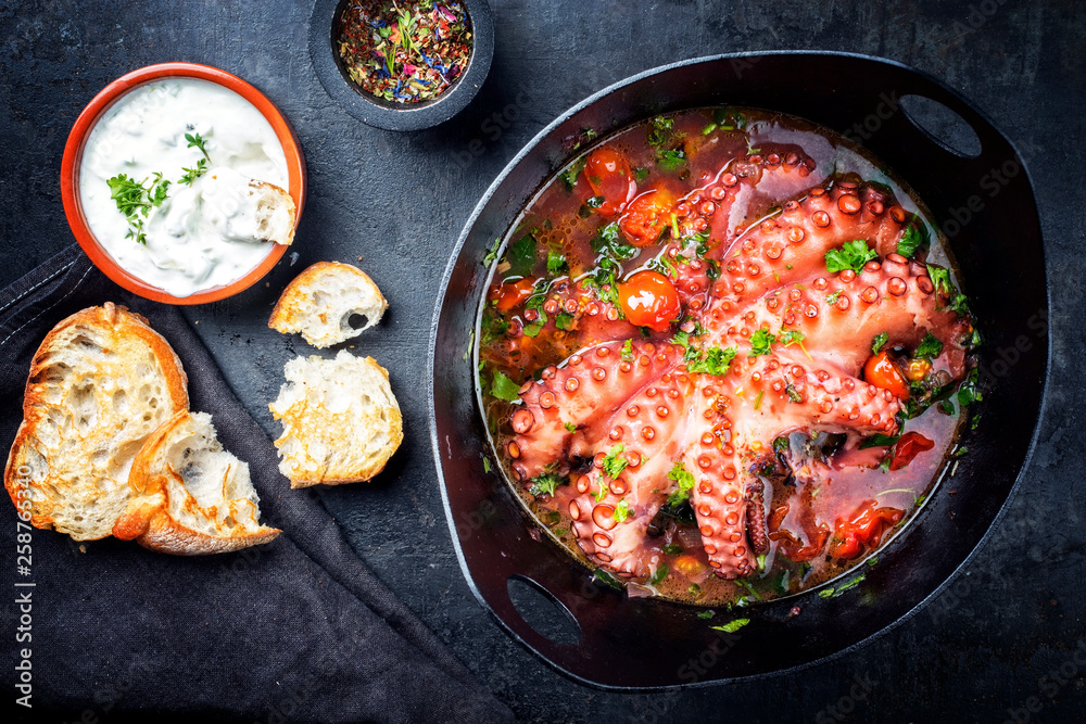 Traditional Greek octopus braised cooked with tomatoes and herbs in ouzo sauce with tzatziki as top view in a cast-iron saucepan