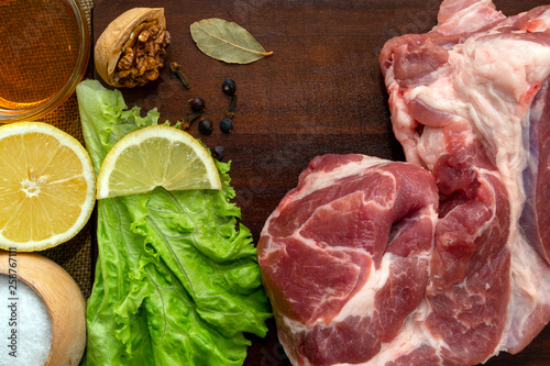 A slice of raw pork meat on a wooden cutting board. Leaf lettuce, seasonings and a slice of lemon. Rustic style, top view.
