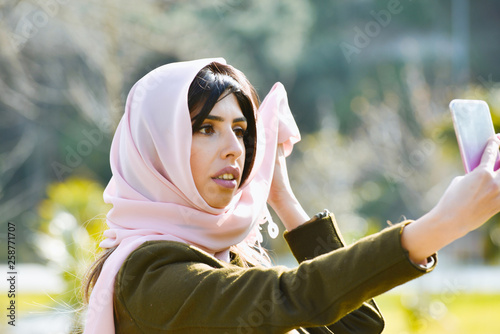 Beautiful young Muslim woman in park smiling at camera, taking selfie